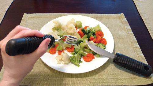 Make Eating Easier Big Grip Utensil Set Blue Jay Brand held by hand with large black ribbed handles cutting and picking vegetables.