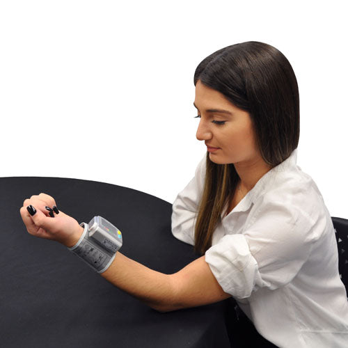 Young woman with dark hair wearing a gray digital wrist cuff blood pressure monitor while seated at black table