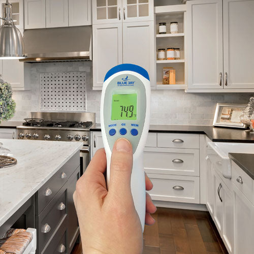 Hand holding white and blue digital thermometer with green screen showing temperature in modern kitchen setting