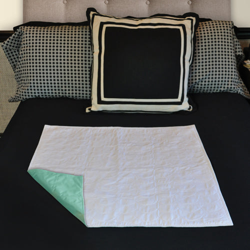 White quilted rectangular pad with light green waterproof backing displayed on black bed with patterned pillows
